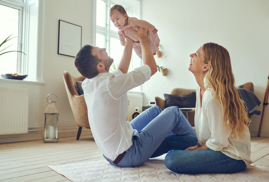 Father holding baby while mother sits on the floor.