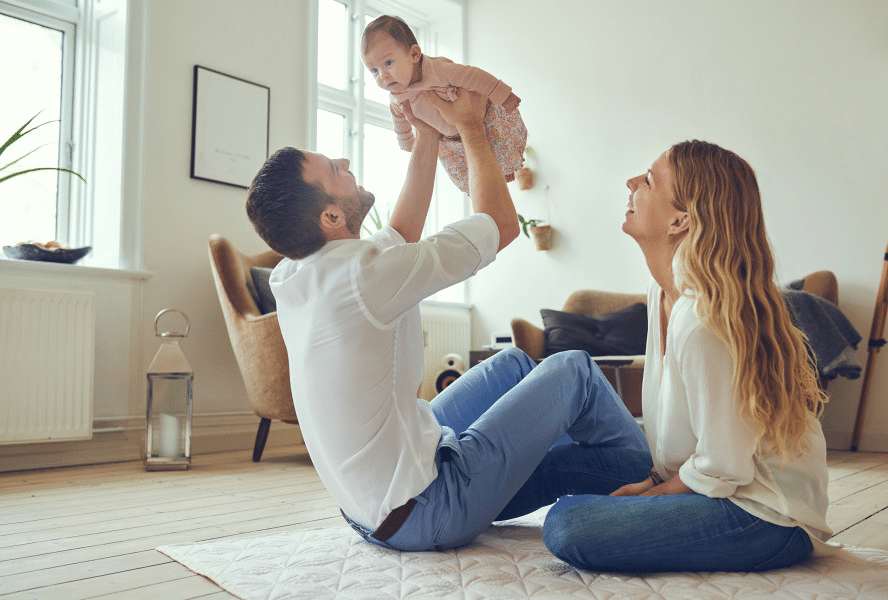 Father holding baby while mother sits on the floor.