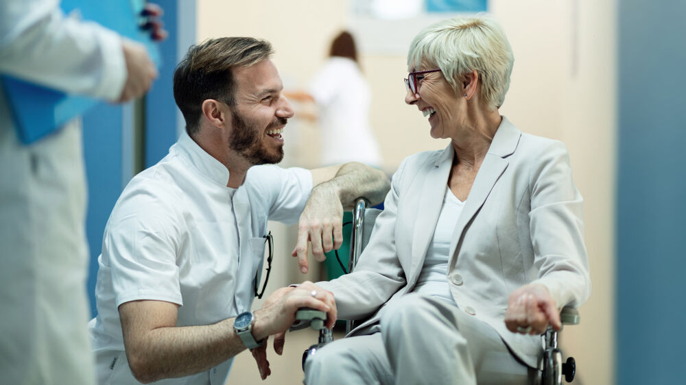 Männlicher Arzt, der sich hinhockt und lächelt, spricht mit einer älteren Frau im Rollstuhl.